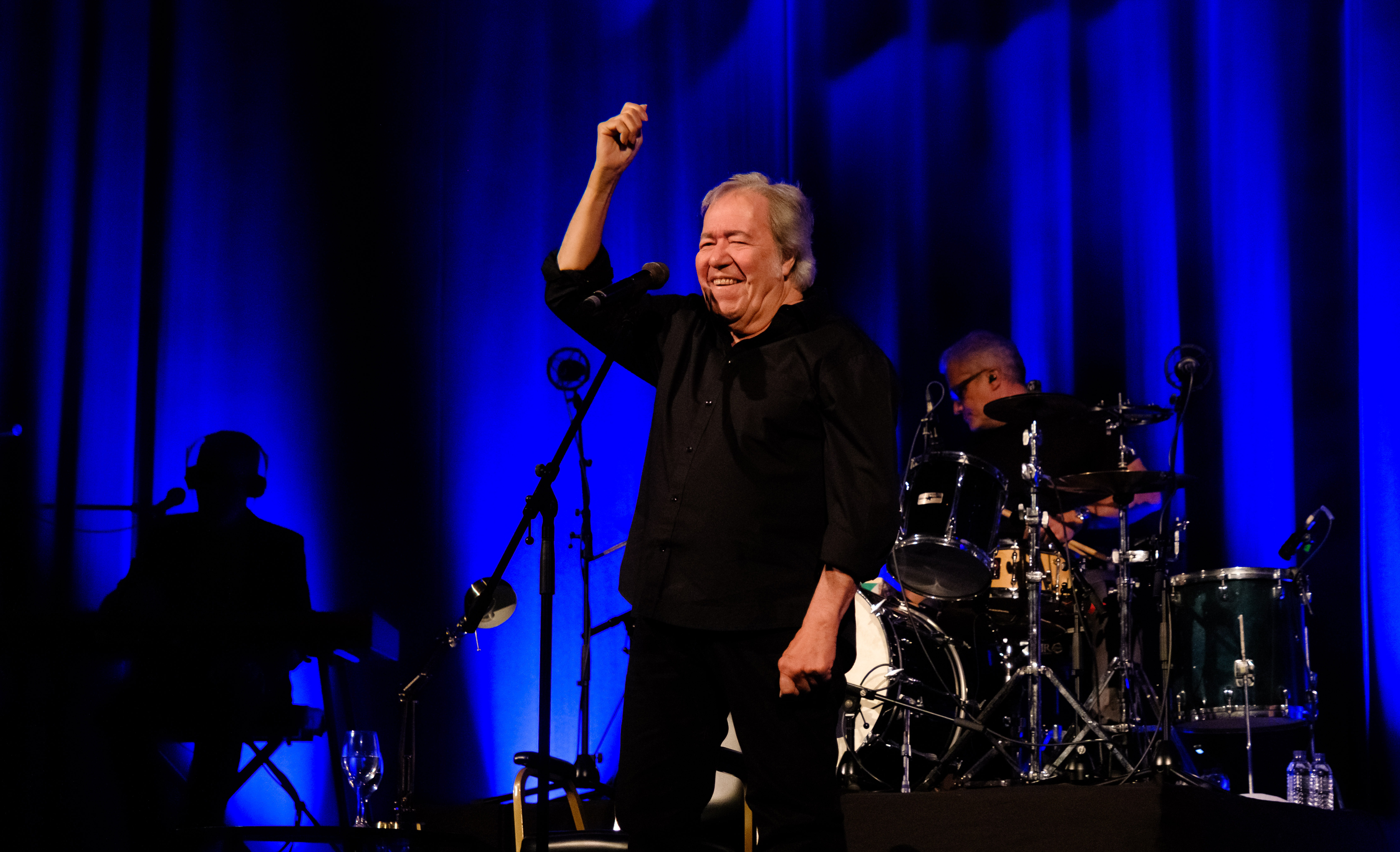 Sérgio Godinho sorridente a cantar no palco com o braço direito levantado