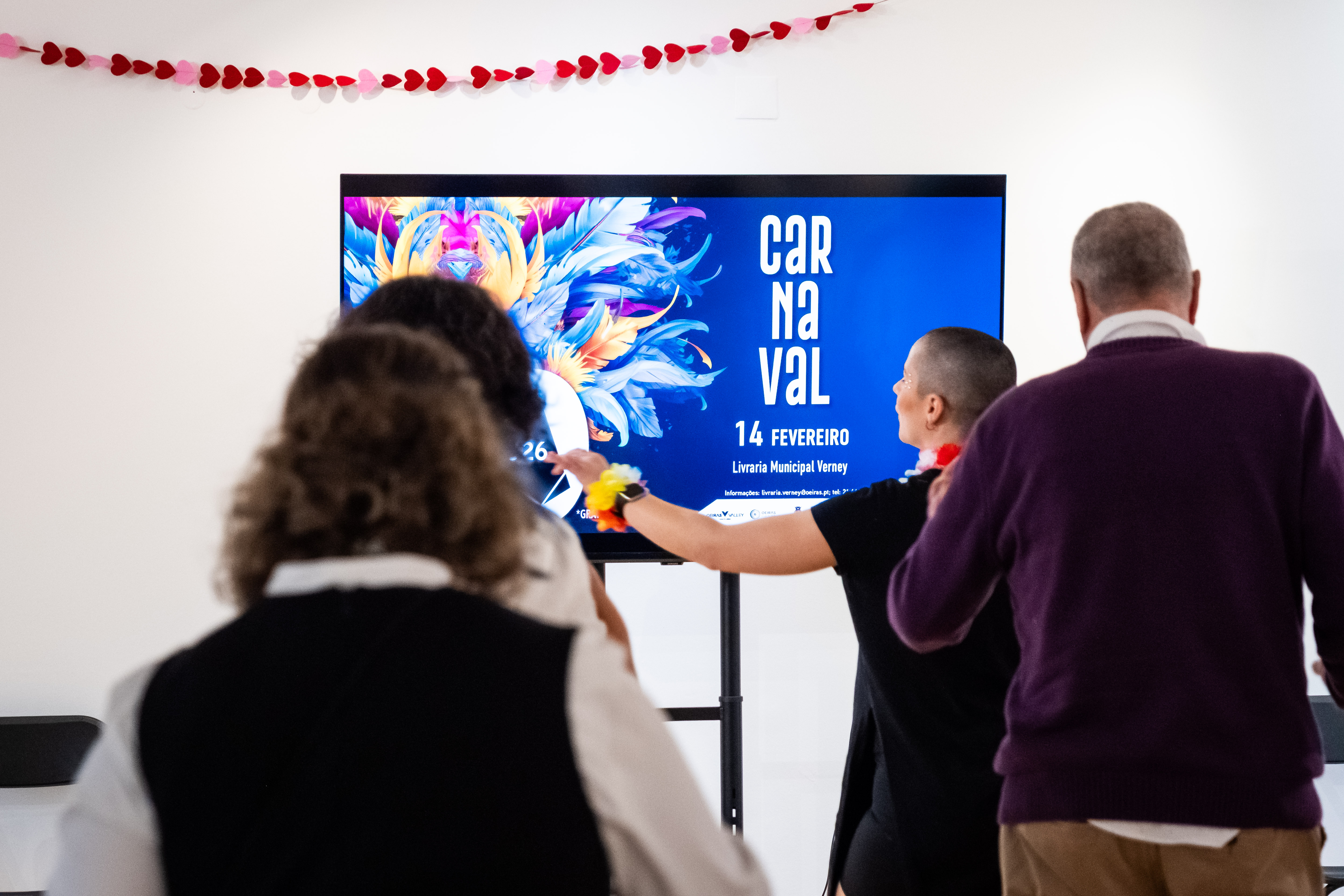 Fotografia com pessoas de costas a dançar e no fundo uma televisão com o banner do evento