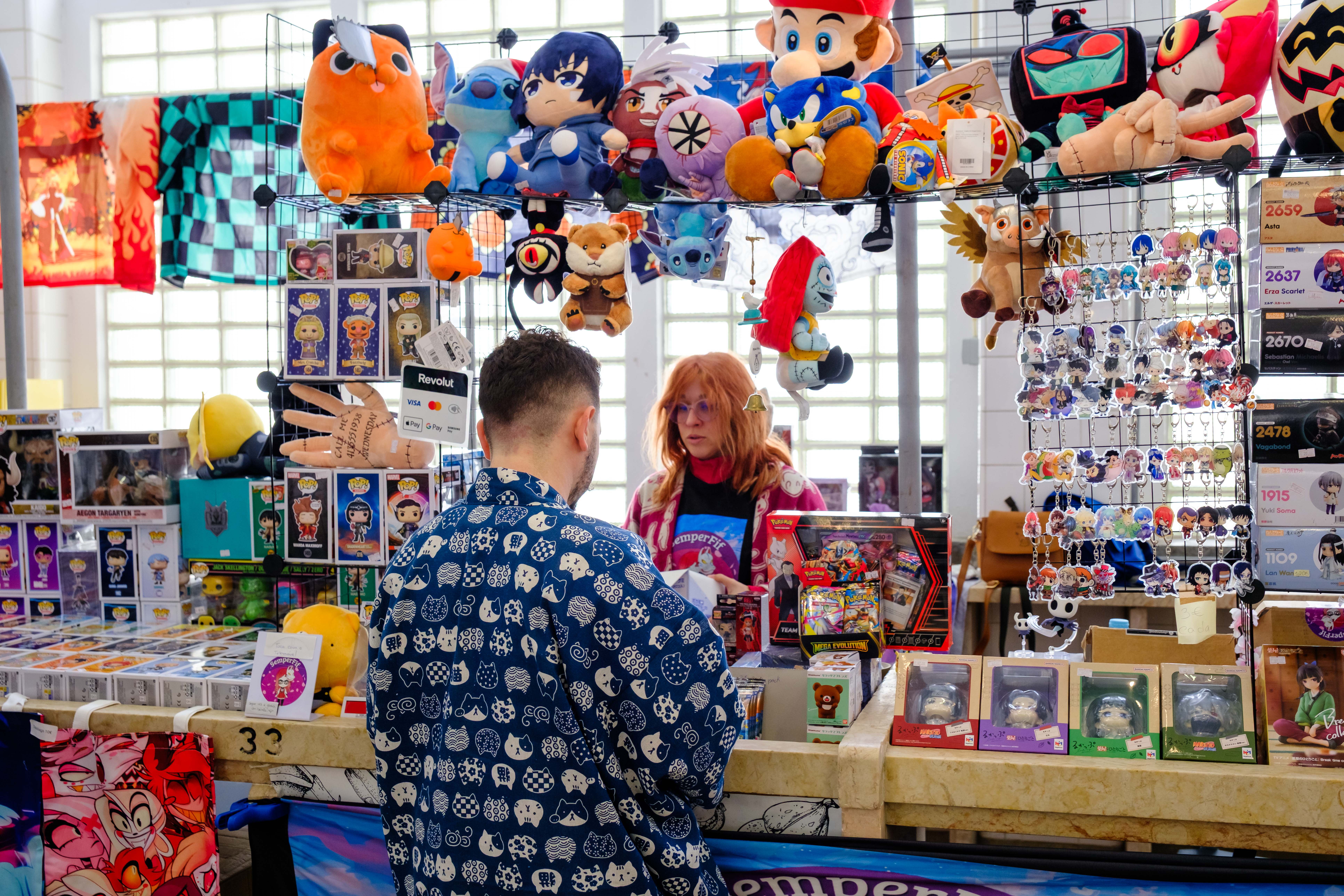 Uma banca com peluches, bonecos e porta-chaves coloridos, onde uma senhora está a atender e um rapaz observa.